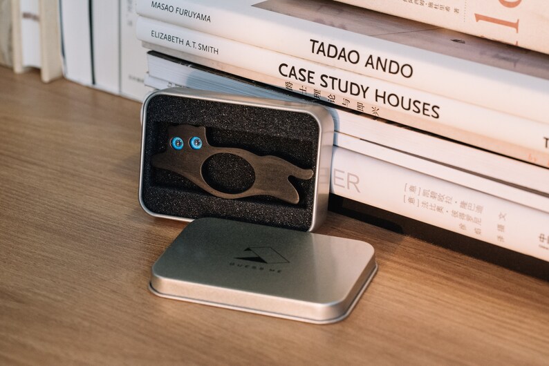 Pu&ograve; includere: Un portachiavi in metallo nero a forma di gatto con occhi blu, in una scatola argentata con interno in schiuma nera. La scatola &egrave; aperta e il portachiavi &egrave; visibile. La scatola &egrave; appoggiata su una superficie di legno.