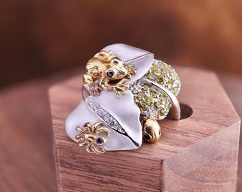 May include: A detailed frog ring with a gold frog, a silver leaf, and a cluster of green gems. The ring also features a small gold spider and is set on a wooden block.