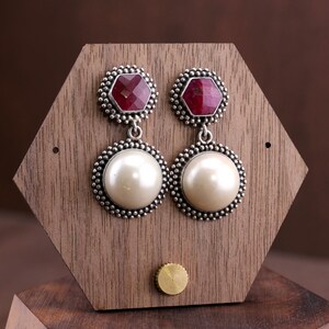 May include: A pair of drop earrings featuring a faceted hexagonal deep red gemstone at the top and a large, smooth, round white pearl at the bottom. Both elements are set in a textured, beaded silver-toned metal frame with a dangling design.