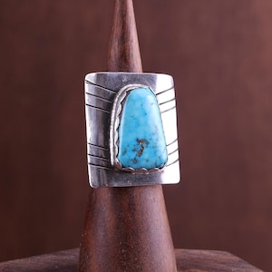 May include: A sterling silver ring featuring a large, trapezoidal turquoise stone with brown matrix inclusions. The silver band has a wide, flat face with etched horizontal line details on both sides of the central stone setting.