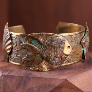 May include: A detailed bronze cuff bracelet featuring an embossed fish design. The open-ended bracelet has a textured surface with an aged patina, showcasing intricate patterns and a fish motif. The bracelet is approximately 2 inches wide.