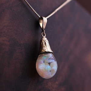 May include: A gold pendant necklace featuring a teardrop-shaped glass vessel filled with iridescent white opal fragments. The pendant is suspended from a delicate gold chain and capped with a decorative gold metal bail with etched detailing.