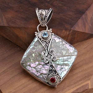 May include: A square-shaped pendant featuring a mosaic of iridescent mother-of-pearl tiles set in silver. The piece is accented with a small round blue gemstone at the top and a round red gemstone at the bottom, connected by silver scrollwork.