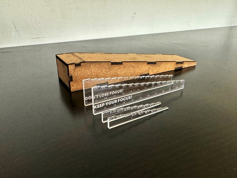 May include: Four clear acrylic rulers with the text "Don't lose focus!" and "Keep your focus!" printed on them. The rulers are arranged in descending order of size on a dark brown surface. A wooden box with a sloped top is in the background.