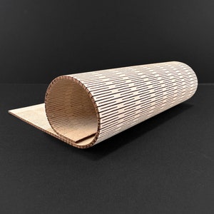 May include: A rolled-up, cylindrical wooden object with a series of evenly spaced, horizontal lines cut into its surface. The object is made of light-colored wood and sits partially unrolled on a flat wooden surface. The background is a solid black.