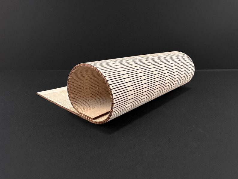 May include: A rolled-up, cylindrical wooden object with a series of evenly spaced, horizontal lines cut into its surface. The object is made of light-colored wood and sits partially unrolled on a flat wooden surface. The background is a solid black.