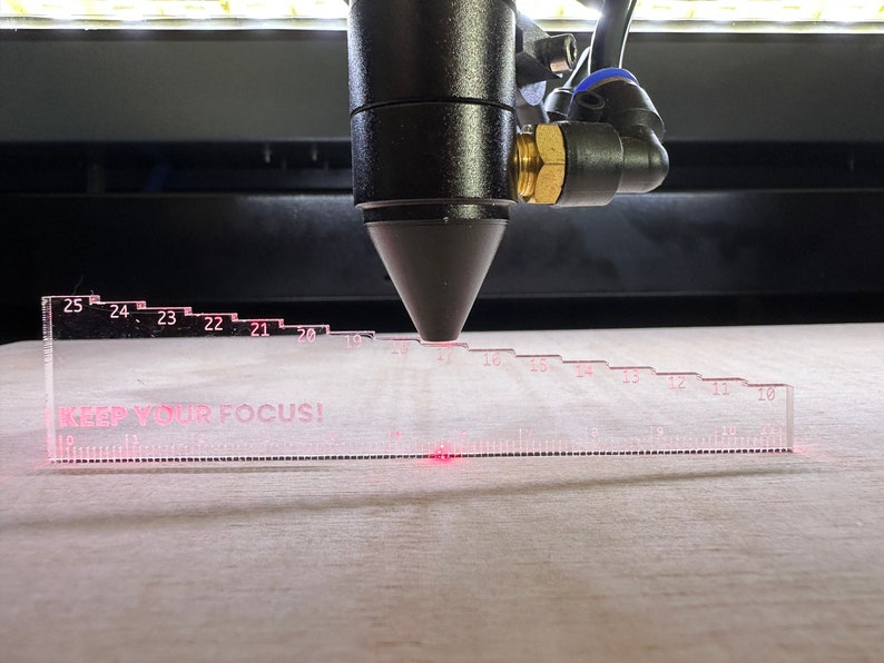 May include: A close-up of a laser cutter head positioned over a clear plastic ruler with the text "KEEP YOUR FOCUS!" printed on it. The ruler has markings from 10 to 25. A red laser beam is visible on the ruler.