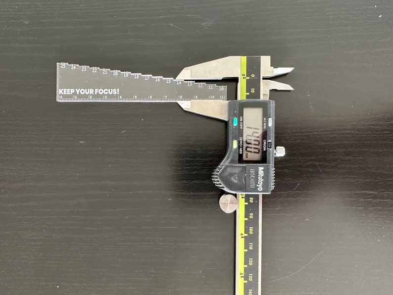 May include: A black and silver digital caliper with a yellow stripe on the ruler. The caliper is open and the display reads 14.00 mm. The ruler has markings in millimeters and inches. The caliper is on a black surface. The ruler has the text "KEEP YOUR FOCUS!" printed on it.