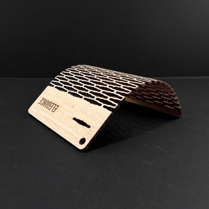 May include: A wooden phone stand with a curved design. The stand is made of light-colored wood with a dark, patterned top section. The word "ELEGANCE" is laser-etched on the base. The stand has a small hole and slots for cable management.