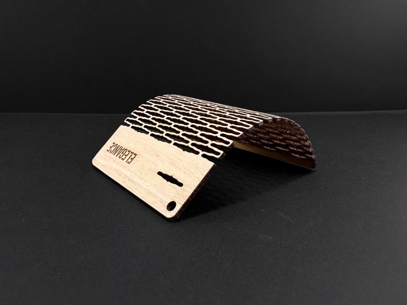 May include: A wooden phone stand with a curved design. The stand is made of light-colored wood with a dark, patterned top section. The word "ELEGANCE" is laser-etched on the base. The stand has a small hole and slots for cable management.