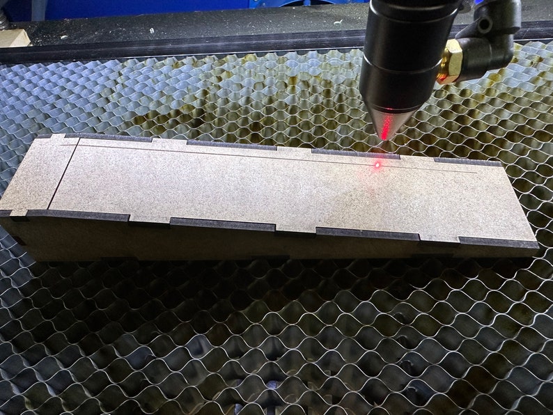 May include: A laser cutter with a red laser beam focused on a piece of wood. The wood is on a honeycomb-patterned surface.