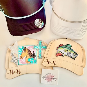 Puede incluir: Dos gorras de camionero, una negra y otra blanca, se muestran sobre dos placas de madera con forma de sombrero. Las placas presentan un caballo y una cruz con detalles turquesas. También hay una pequeña tarjeta con el texto "Hatzel & Harmony".