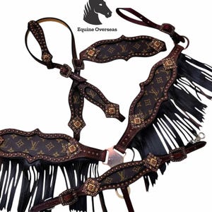 Op de afbeelding: Een bruine en tan paardentrenssset met een herhalend patroon en goudkleurige accenten. De trens heeft zwarte franjes en decoratieve metalen hardware. De merknaam "Equine Overseas" is zichtbaar.