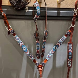 Pode incluir: Um conjunto de arreios para cavalos com peitoral, cabeçada e rédeas. O conjunto é feito de couro marrom com intrincados trabalhos de contas em padrões geométricos turquesa, vermelhos e brancos. Ferragens prateadas acentuam as peças. Uma corda de couro enrolada também é visível.