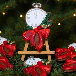 Può includere: Biscotti natalizi a forma di orologi da tasca e fiocchi. I biscotti sono decorati con fiocchi rossi, rametti verdi e dettagli dorati. Un biscotto è esposto su un piccolo cavalletto di legno. Lo sfondo è un albero di Natale con luci scintillanti.