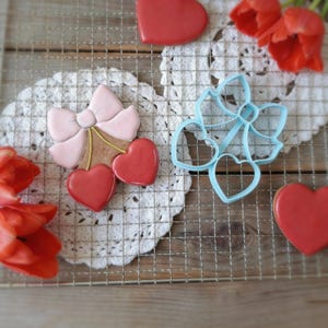 Puede incluir: Una galleta decorada con forma de lazo y dos cerezas, con un lazo rosa claro y cerezas rojas. Un cortador de galletas azul en forma de corazón está al lado de la galleta. Galletas rojas en forma de corazón y tulipanes rojos también están en la imagen.