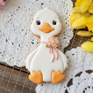 Può includere: Un biscotto bianco a forma di oca con zampe e becco arancioni, ornato da un fiore e un nastro rosa. Il biscotto è appoggiato su una tovaglietta bianca, con narcisi gialli e fiori rosa sullo sfondo. Una delizia affascinante.