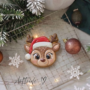 May include: A decorated cookie shaped like a reindeer head wearing a red and white Santa hat with brown antlers, large eyes, and a small nose, positioned on a wire cooling rack alongside bronze-colored ornaments and white snowflake decorations.