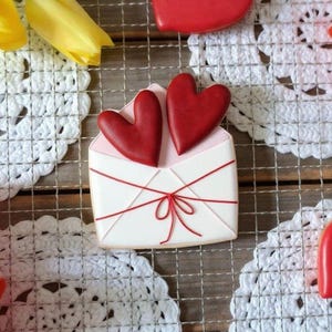 Könnte beinhalten: Eine Anordnung von Keksen zum Valentinstag. Ein weißer Keks in Umschlagform mit roten Schnurdetails und zwei roten Herz-Keksen. Gelbe Tulpen und weitere Herz-Kekse sind ebenfalls zu sehen.