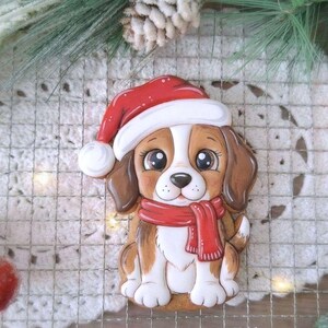 May include: A festive dog-shaped cookie, decorated with brown, white, and red icing. The dog wears a Santa hat and a red scarf. The cookie is placed on a white, patterned surface, with Christmas decorations in the background.