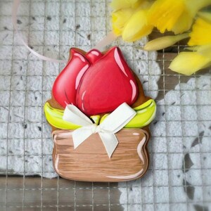 Peut inclure: Ornement décoratif en forme de tulipe en bois. La tulipe est rouge avec une base marron et des accents jaunes. Un ruban blanc est noué autour de la base. L'ornement est sur une surface en dentelle blanche avec des fleurs jaunes.