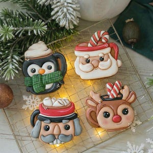 Op de afbeelding: Vier feestelijke kerstkoekjes in de vorm van mokken. Ontwerpen omvatten een pinguïn met een sjaal, de kerstman, een rendier en een notenkraker. De koekjes zijn versierd met glazuur en geplaatst op een draadrek met kerstversieringen.