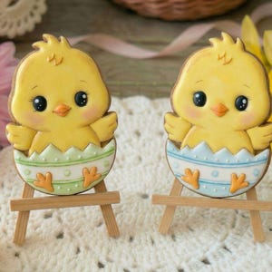 Könnte beinhalten: Zwei gelbe Küchlein in Form von Küken, jeweils in einer verzierten Eierschale, stehen auf kleinen Holzstaffeleien. Die Küchlein haben schwarze Augen, orangefarbene Schnäbel und sind mit Pastellfarben verziert. Der Hintergrund umfasst Blumen und eine Spitzendecke.
