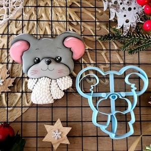 Peut inclure: Un biscuit en forme de souris grise avec des oreilles roses et une écharpe blanche, à côté d'un emporte-pièce bleu de la même forme. Le biscuit a des yeux noirs et des moustaches. Éléments décoratifs : flocons de neige en bois et baies rouges.