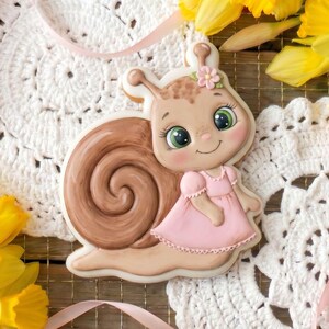 Könnte beinhalten: Ein dekorativer Keks in Schneckenform mit braunem Gehäuse und rosa Kleid. Der Keks hat ein lächelndes Gesicht mit grünen Augen und einer rosa Blume. Umgeben von gelben Narzissen und rosa Blumen, auf einer weißen Spitze.