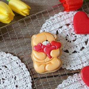 Op de afbeelding: Een koekje in de vorm van een teddybeer met rode hartvormige snoepjes. De koek is lichtbruin met een wit en zwart gezicht. Rode hartsnoepjes en gele tulpen staan ook op de foto.