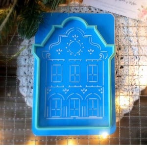 Può includere: Un tagliabiscotti a forma di casa blu con dettagli bianchi. Il tagliabiscotti presenta una facciata dettagliata con finestre e porte. Il tagliabiscotti è realizzato in plastica ed è progettato per la cottura. L'immagine include anche decorazioni natalizie e verde.