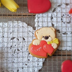 Könnte beinhalten: Ein Teddybär-Keks, der zwei rote herzförmige Kekse hält, von denen einer die Aufschrift "I love you" trägt. Der Bär hat eine gelbe Fliege. Gelbe Tulpen und rote herzförmige Kekse sind im Hintergrund.