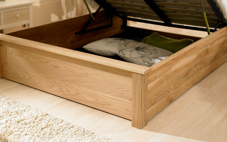 May include: A wooden bed frame with a lift-up storage compartment. The compartment is open and shows a white and floral patterned pillow and a green pillow.