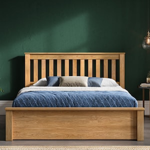 May include: A wooden bed frame with a slatted headboard and footboard. The bed is made of natural oak wood and has a storage drawer underneath. The bed is set against a dark green wall and has a blue blanket and pillows on top.