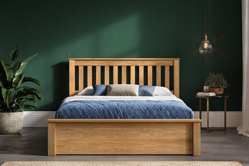 May include: A wooden bed frame with a slatted headboard and footboard. The bed is made of natural oak wood and has a storage drawer underneath. The bed is set against a dark green wall and has a blue blanket and pillows on top.