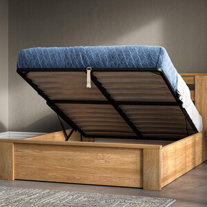 May include: A light wood bed frame with a blue upholstered headboard and a lift-up storage compartment. The bed frame is open and the mattress is lifted up to reveal the storage space.