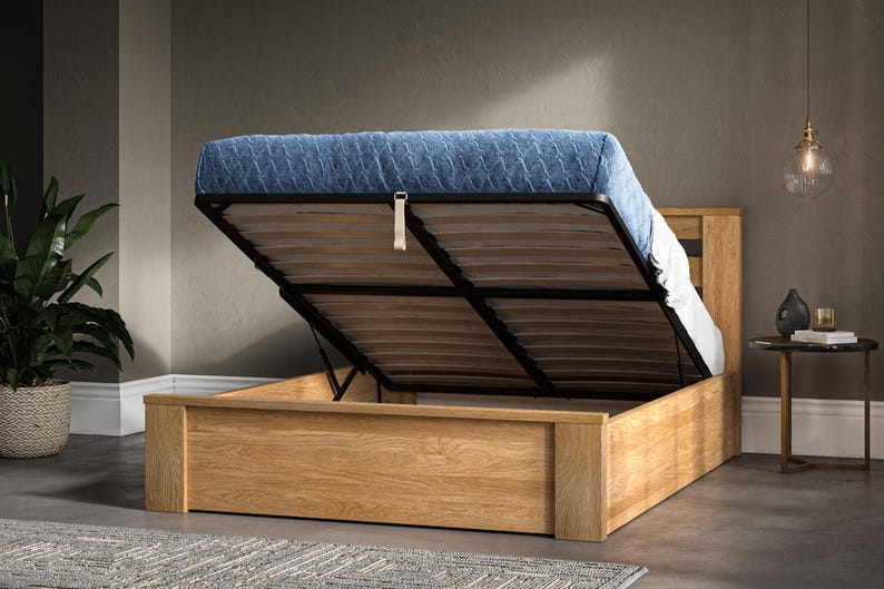 May include: A light wood bed frame with a blue upholstered headboard and a lift-up storage compartment. The bed frame is open and the mattress is lifted up to reveal the storage space.