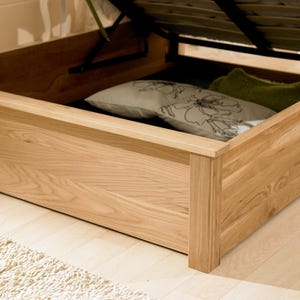 May include: A wooden bed frame with a lift-up storage compartment. The compartment is open and shows a white and floral patterned pillow and a green pillow.