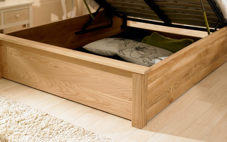May include: A wooden bed frame with a lift-up storage compartment. The compartment is open and shows a white and floral patterned pillow and a green pillow.