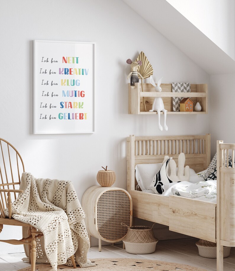 May include: A white framed print with colourful text in German that reads "Ich bin" followed by a list of positive adjectives: nett, kreativ, klug, mutig, stark, geliebt. The print is hanging on a white wall in a child's bedroom.