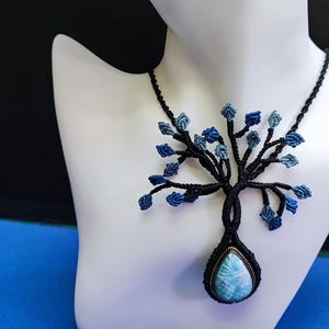 May include: A macrame tree of life necklace with a black cord and blue accents. The pendant features a teardrop-shaped, light blue gemstone. The necklace is displayed on a white mannequin.