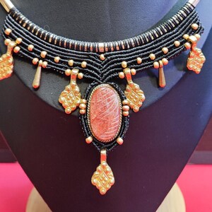May include: A handcrafted necklace featuring a black macrame design with gold-colored accents. The focal point is an oval, reddish-brown stone pendant. The necklace is displayed on a black bust.
