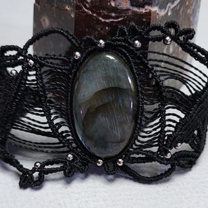 May include: Black macramé bracelet with an oval labradorite stone. The bracelet has a woven design with small silver beads. The background features a natural stone with white and brown patterns.