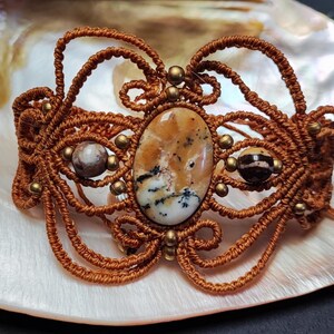 Peut inclure: Un bracelet macramé fait à la main avec une pierre centrale ovale aux motifs marron, beige et noirs. Le bracelet est fait de cordon marron et orné de petites perles dorées. Le bracelet est présenté sur une surface blanche.