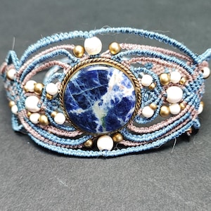 Op de afbeelding: Handgemaakte macram&eacute; armband met een centrale blauwe sodalietsteen in een messing zetting. De armband gebruikt blauwe en bruine koorden, met witte en gouden kralen. Een uniek sieraad.