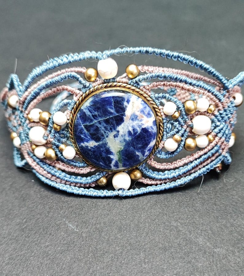 Op de afbeelding: Handgemaakte macram&eacute; armband met een centrale blauwe sodalietsteen in een messing zetting. De armband gebruikt blauwe en bruine koorden, met witte en gouden kralen. Een uniek sieraad.