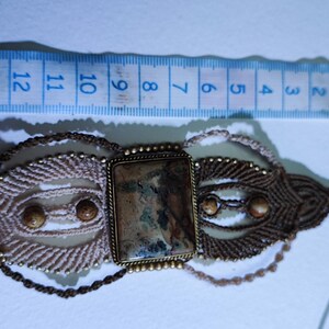 May include: A handmade macrame choker necklace with a rectangular stone centerpiece. The necklace features a beige and brown woven design with small, round beads. A blue ruler is placed next to the necklace, showing measurements in centimeters.