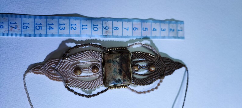 May include: A handmade macrame choker necklace with a rectangular stone centerpiece. The necklace features a beige and brown woven design with small, round beads. A blue ruler is placed next to the necklace, showing measurements in centimeters.
