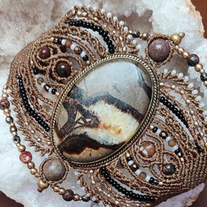 May include: A macrame bracelet featuring a large oval brown and cream patterned stone set in a brass-toned metal frame. The band is crafted from brown cord with intricate knotting, accented by small black, white, and metallic beads and round stones.
