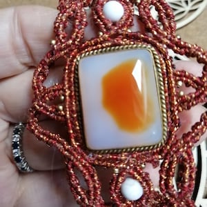Può includere: Un braccialetto in macramé rosso e oro con una pietra di agata arancione quadrata al centro. Il braccialetto è decorato con perline bianche.